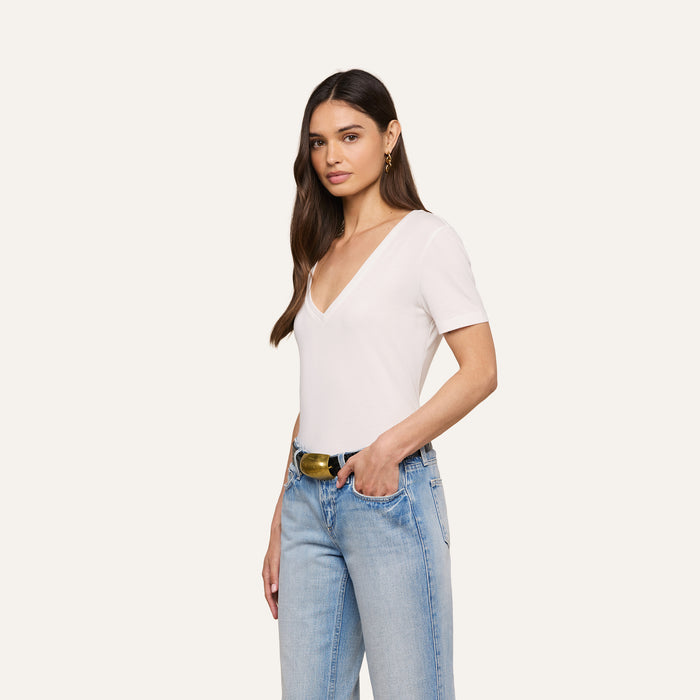 A woman with long brown hair stands against a plain light background with one hand in her pocket, wearing the relaxed fit Brianna Cotton V-Neck Tee, light blue jeans, and a black belt with a gold buckle.