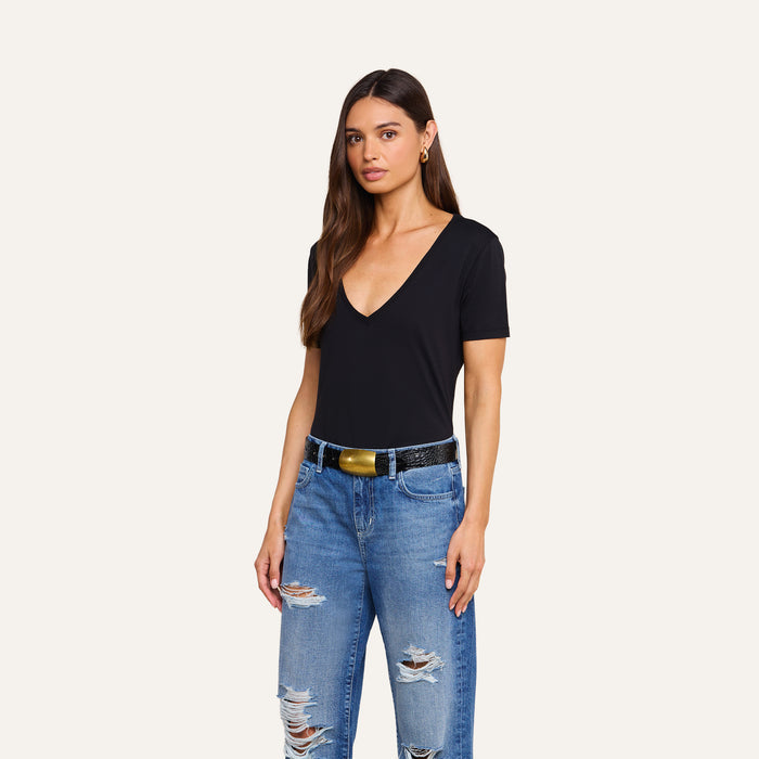 A woman with long brown hair is wearing the Brianna Cotton V-Neck Tee in black—crafted from soft Supima cotton—paired with ripped blue jeans and a black belt with a gold buckle, standing against a plain light background.