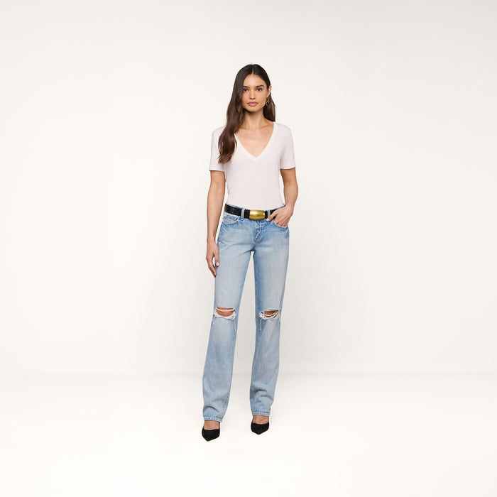 A woman with long brown hair and a neutral expression stands against a plain background, wearing the Brianna Cotton V-Neck Tee—a relaxed-fit white T-shirt—paired with light blue ripped jeans, a black belt, and black heels.
