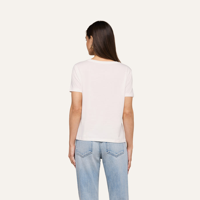A person with long brown hair is shown from the back, standing against a plain light background and wearing the Brianna Cotton V-Neck Tee in white in a relaxed fit, paired with light blue jeans.