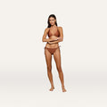 Wearing the Britt Tie-Side Bikini Bottom in bronze, a woman stands barefoot against a plain white background, arms gently crossed and a slight smile on her face.