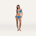 A woman with straight brown hair and a relaxed expression stands barefoot on a plain background, wearing the Britt Tie-Side Bikini Bottom—a capri blue style featuring adjustable tie sides. She pairs it with a matching halter-style top that offers moderate coverage.