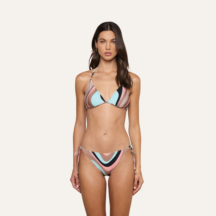 A woman with long brown hair and a neutral expression stands facing forward against a plain light background, wearing the Britt Tie-Side Bikini Bottom in an abstract swirl print of pink, blue, black, and white.