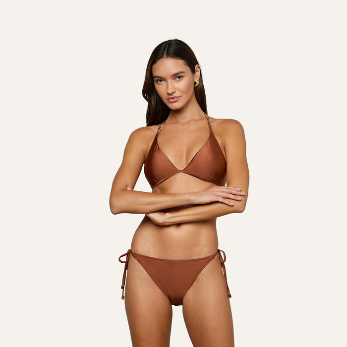A woman stands with her arms crossed over her midsection, modeling the Britt Tie-Side Bikini Bottom in a bronze hue, featuring moderate coverage and adjustable tie sides, set against a plain light background.