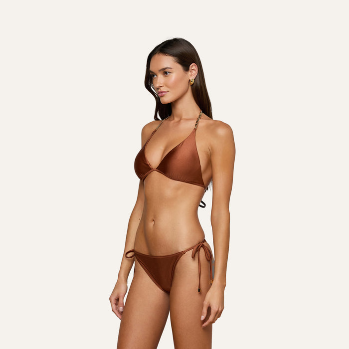 A woman wears the Britt Tie-Side Bikini Bottom in bronze, featuring moderate coverage and adjustable tie sides, as she stands against a plain light background looking slightly to her left with her arms relaxed by her sides.