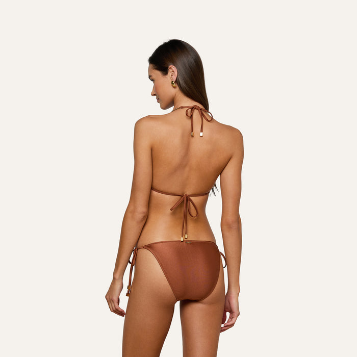 A woman with long brown hair, facing away, is wearing a bronze bikini halter top and the Britt Tie-Side Bikini Bottoms featuring adjustable side ties and moderate coverage, standing against a plain light background.