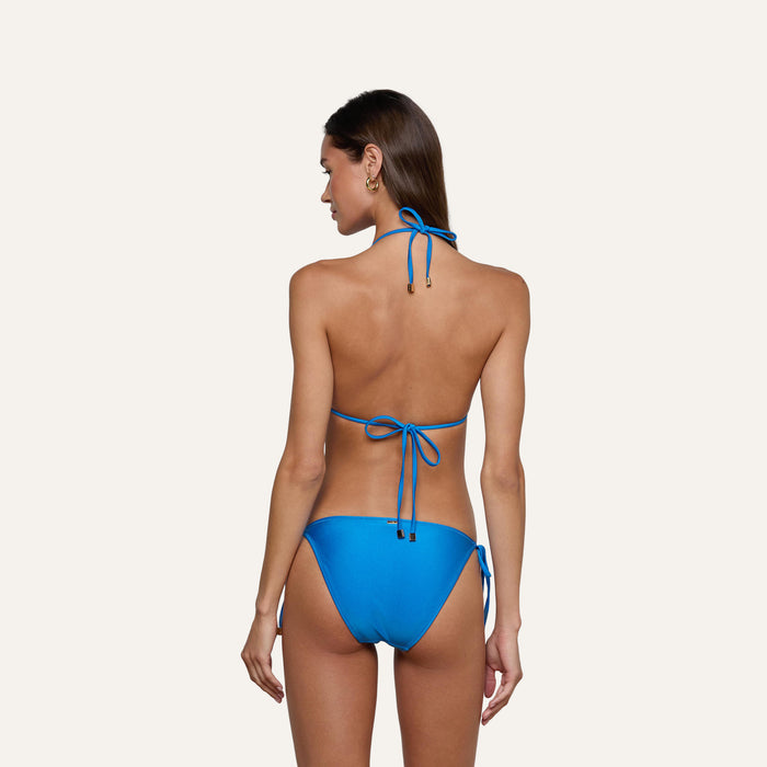 A woman stands with her back to the camera, wearing the Britt Tie-Side Bikini Bottom in capri blue, featuring moderate coverage and adjustable tie sides, set against a plain light background.