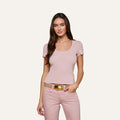 A woman stands against a plain background, wearing the Brynlee Scoopneck Tee in light pink with short sleeves, paired with light pink pants and a beige belt featuring a metallic buckle. She has long brown hair and is smiling slightly.
