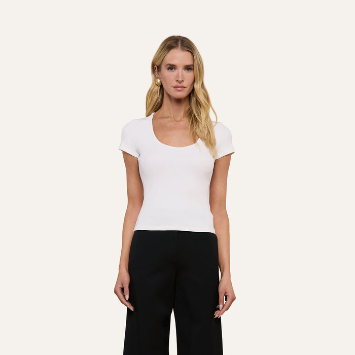 A woman stands against a plain light background wearing the Brynlee Scoopneck Tee—a fitted, white ribbed top with a scoop neckline—paired with black pants. Her arms rest naturally at her sides and her long blonde hair is styled loosely.