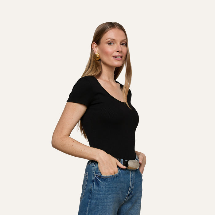 Woman with long, light brown hair wearing the Brynlee Scoopneck Tee in black and blue jeans, accessorized with gold hoop earrings, stands with one hand in her pocket, smiling slightly against a plain light background.