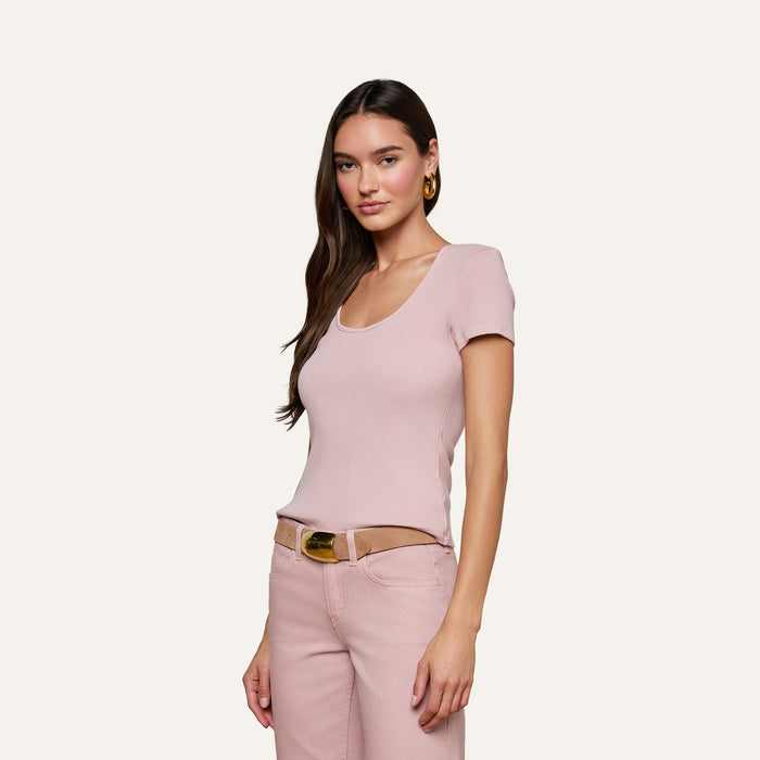 A woman with long dark hair wears the Brynlee Scoopneck Tee in light pink, paired with pink pants and a brown belt. She stands with one hand in her pocket against an off-white background, smiling slightly.