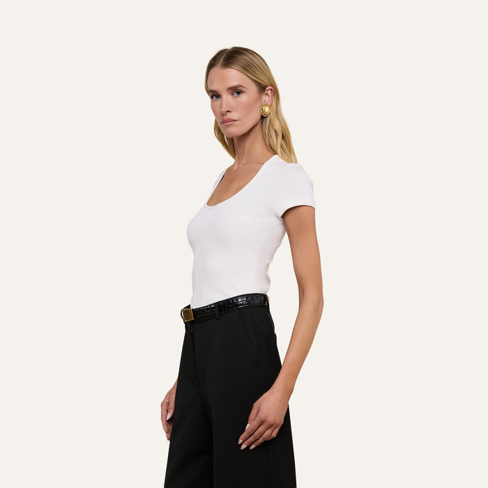 A woman with long blonde hair wears the Brynlee Scoopneck Tee—a fitted white ribbed top—paired with high-waisted black pants, a black belt, and gold earrings, standing against an off-white background.