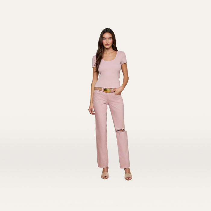 A woman with long brown hair poses in a relaxed stance against a white background, wearing the Brynlee Scoopneck Tee in light pink—crafted from stretch cotton-modal—paired with pink pants featuring a rip at one knee, nude heels, and a yellow belt.