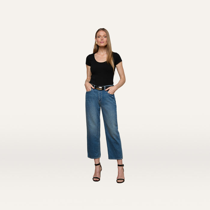 A woman stands against a plain white background, wearing the Brynlee Scoopneck Tee in black, paired with blue high-waisted jeans, a black belt, and black high-heeled sandals, with her hands in her pockets.