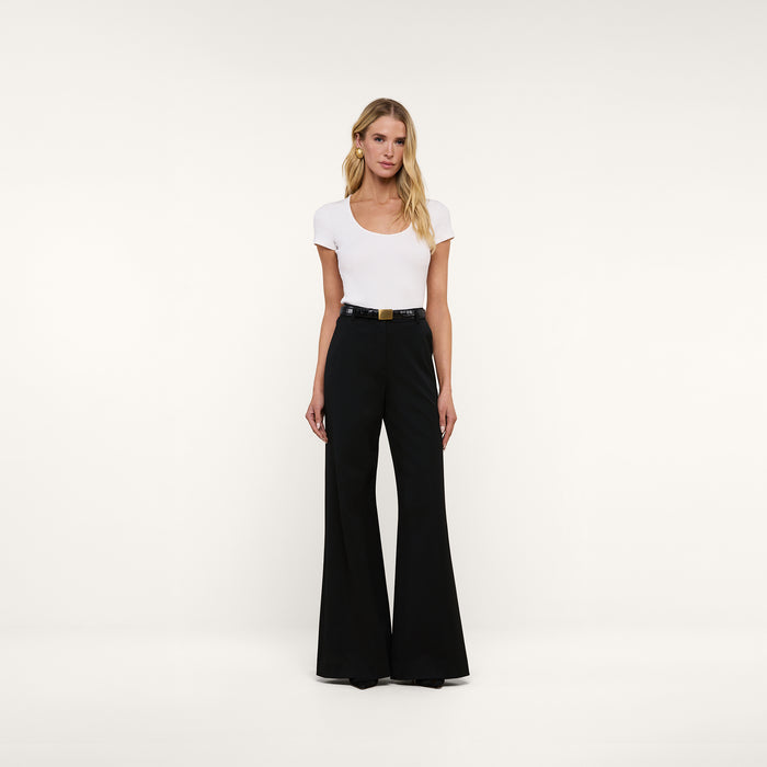 A woman stands against a plain white background, wearing the Brynlee Scoopneck Tee—a fitted, ribbed stretch cotton short-sleeve white top—tucked into high-waisted, wide-leg black pants with a belt, and paired with black shoes.