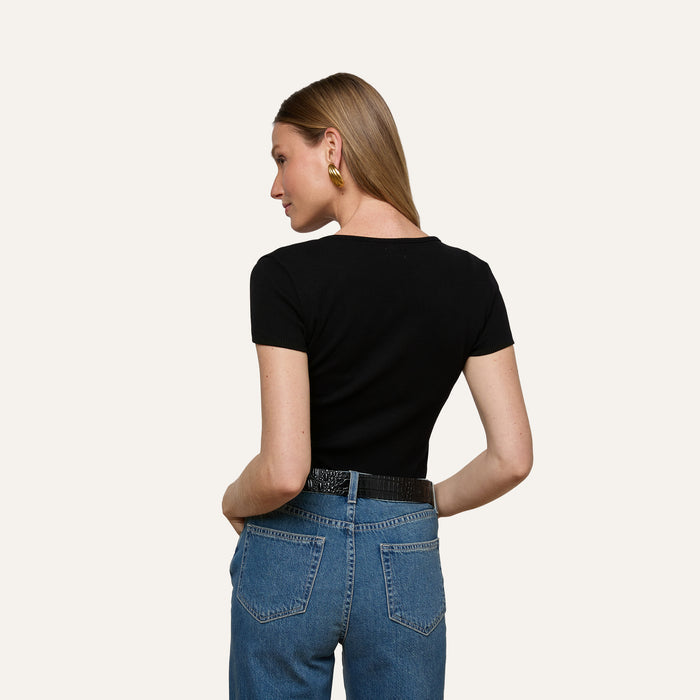 A woman with straight light brown hair, wearing the Brynlee Scoopneck Tee in black with short sleeves, blue jeans, gold earrings, and a black belt, stands with her back to the camera and her hands in her back pockets.