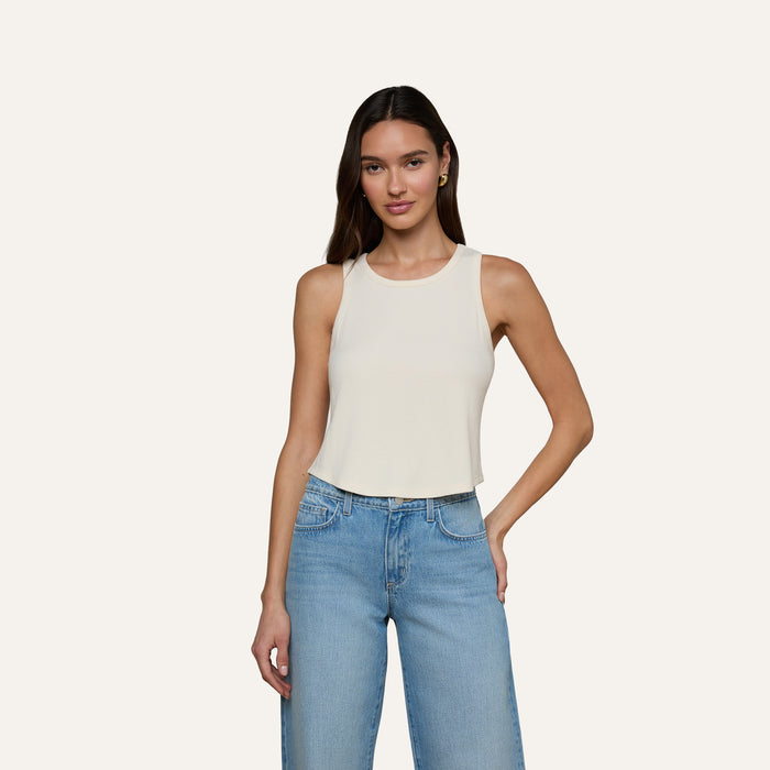 A woman stands against a plain off-white background, wearing the Cael Tank Top—an ultra-soft, sleeveless white crop top—paired with light blue jeans and her right hand in her pocket.