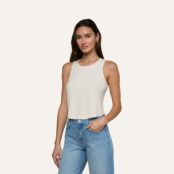 A woman wearing the Cael Tank Top in white and light blue jeans stands against a plain light background, one hand in her pocket and a slight smile.