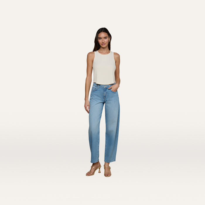 A woman stands against a plain white background, wearing the Cael Tank Top in white, paired with light blue high-waisted jeans and tan heeled sandals. She has long hair and poses relaxedly with one hand in her pocket.