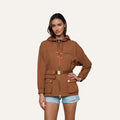 Wearing the Calina Windbreaker—a brown, utility-chic hooded jacket with gold zippers and buttons and a belted waist—she pairs it with light blue distressed denim shorts, posing against a plain light background.