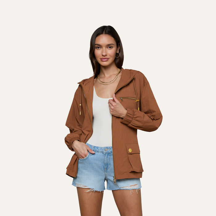 A woman stands against a plain light background with one hand in her pocket, sporting the Calina Windbreaker in brown over a white top and light blue denim shorts for a chic utility-inspired look.