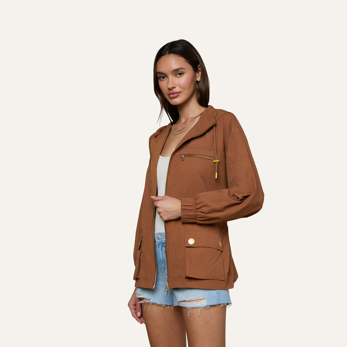 A woman stands against a plain light background, wearing the Calina Windbreaker in brown over a white top and distressed denim shorts.