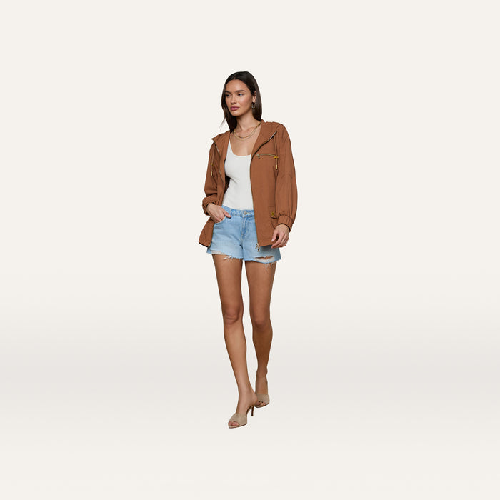 A woman stands against a plain white background wearing the Calina Windbreaker in a utility-chic brown, styled with a white tank top, denim shorts, and beige heels.