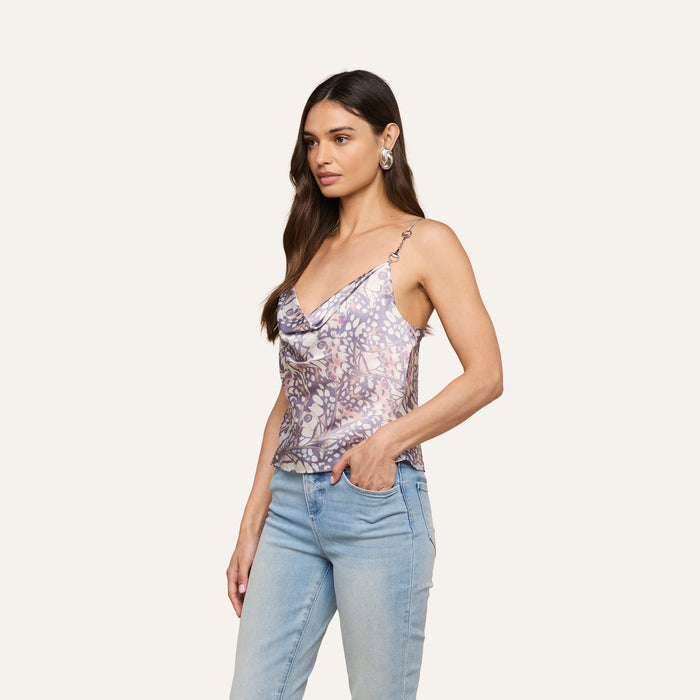 Woman with long brown hair wearing the Calista Satin Camisole—a floral-patterned, bias-cut satin sleeveless cowl-neck top—paired with light blue jeans, standing with one hand in her pocket against a plain background.