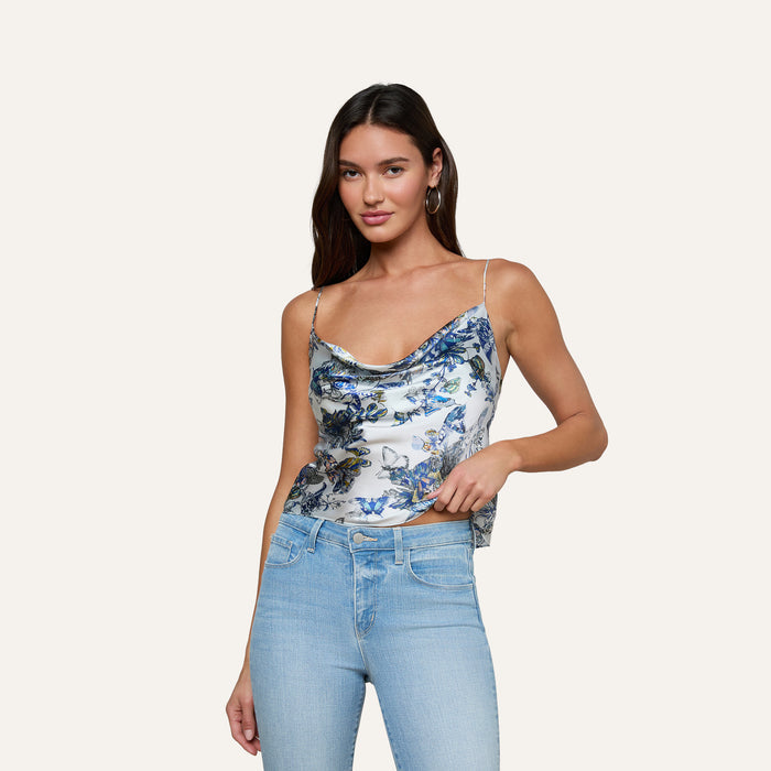 A woman wearing the Calista Silk Camisole with a blue butterfly print and light blue jeans stands against a plain, light background, holding the edge of her top with one hand while the other rests by her side.