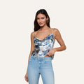 A woman wearing the Calista Silk Camisole with a blue butterfly print and light blue jeans stands against a plain, light background, holding the edge of her top with one hand while the other rests by her side.