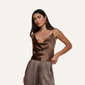 A woman stands against a plain light background wearing the Calista Silk Camisole in brown, styled with high-waisted, pleated brown pants. She has long, straight dark hair and accessorizes with gold jewelry.
