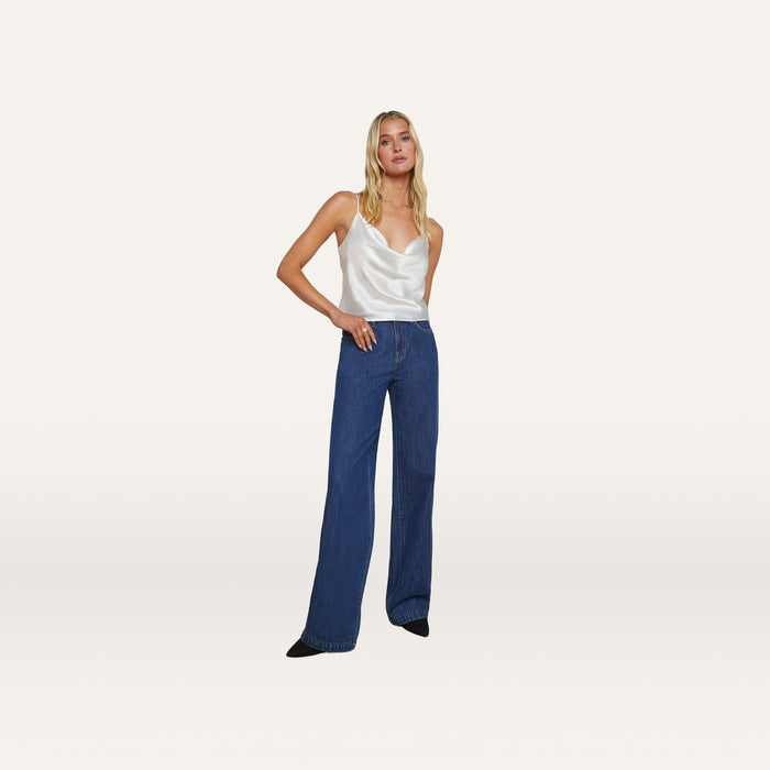 A woman stands against a plain background, wearing the Calista Silk Camisole—crafted from white silk charmeuse with a weighted cowl neck—paired with high-waisted, wide-leg blue jeans and black pointed shoes.