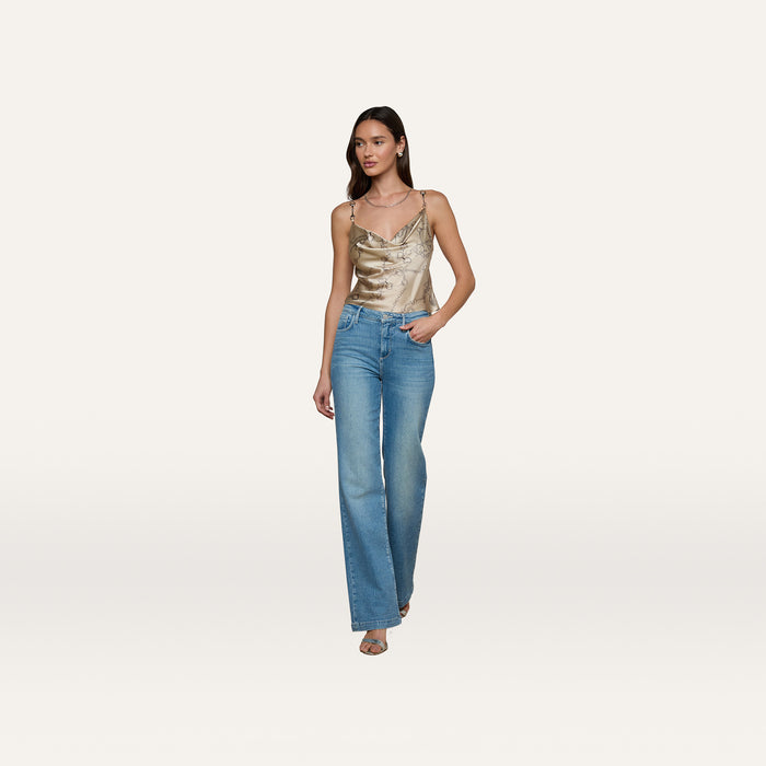 A woman poses against a plain, light background wearing the Calista Silk Camisole in shiny gold, paired with high-waisted blue jeans and heeled sandals.