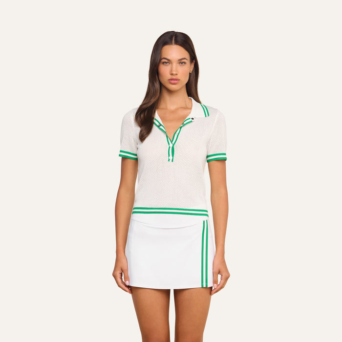 A woman stands facing forward, wearing the Calvin Polo Top—a featherweight white knit short-sleeve tennis polo shirt with green trim—paired with a white skirt featuring green stripes on the side, against a plain light background.