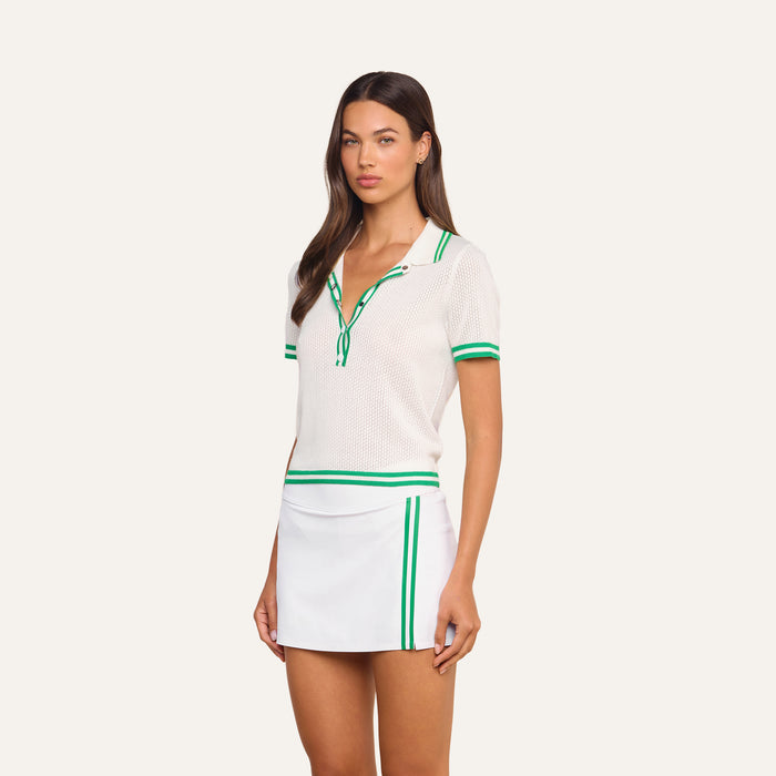 A woman stands against a plain background wearing the Calvin Polo Top, a featherweight knit tennis polo with green stripes, paired with a white mini skirt featuring green accents.
