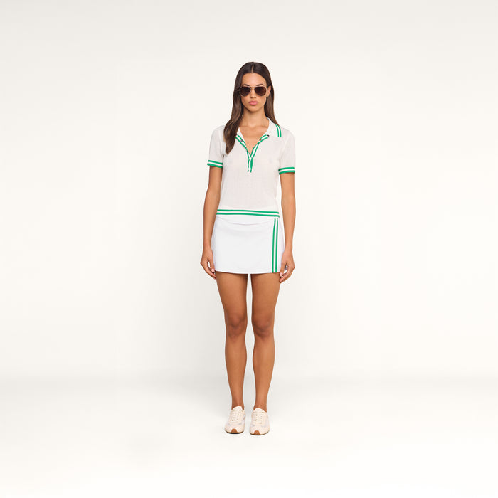 A woman wearing sunglasses, the Calvin Polo Top—a featherweight knit tennis polo with green trim—and a white skirt with green stripes stands facing forward against a plain white background.