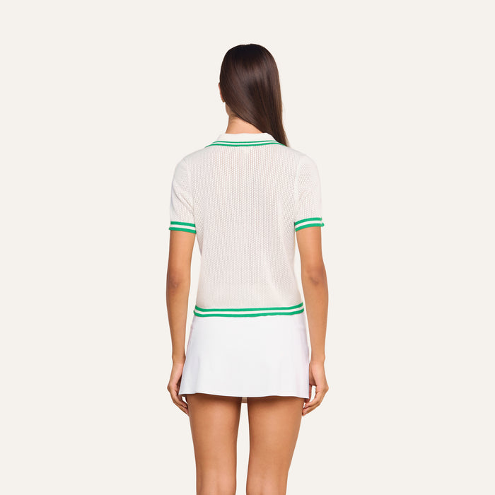 A woman stands with her back to the camera, wearing the Calvin Polo Top—a white short-sleeve tennis polo with green trim—paired with a white skirt against a plain, light-colored background.
