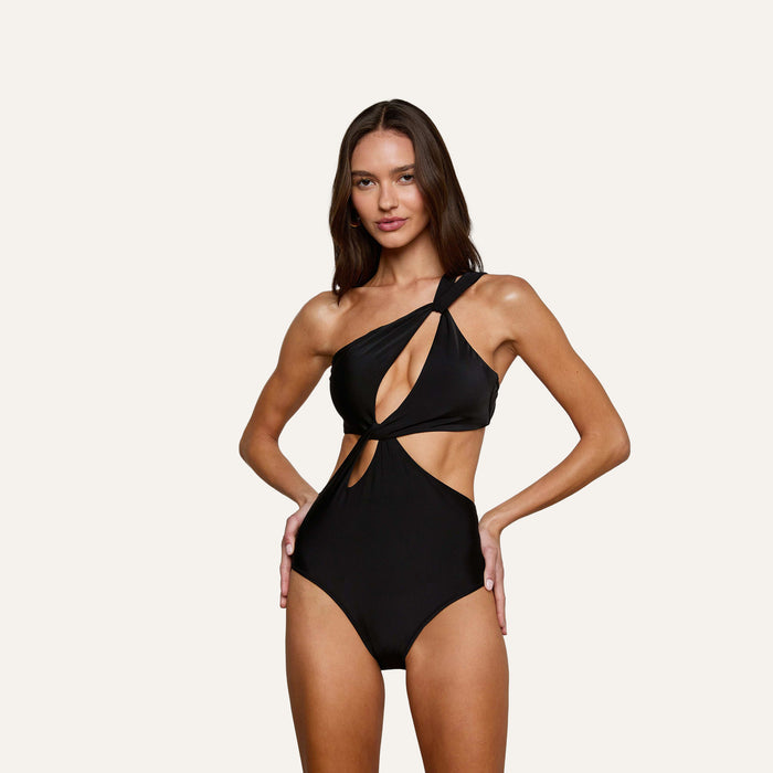 A woman models the Camilla Monokini—a black, one-shoulder swimsuit with a cutout design—posing with her hands on her hips and smiling against a plain light background.