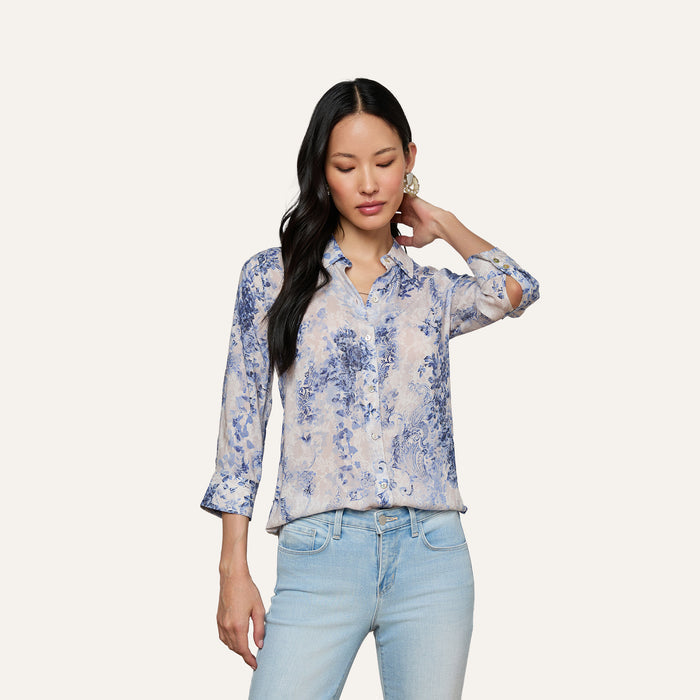 A woman stands against a plain background, looking down with one hand touching her hair, wearing the Camille Blouse—an ivory and blue lace print three-quarter sleeve shirt made from viscose twill—paired with light blue jeans.