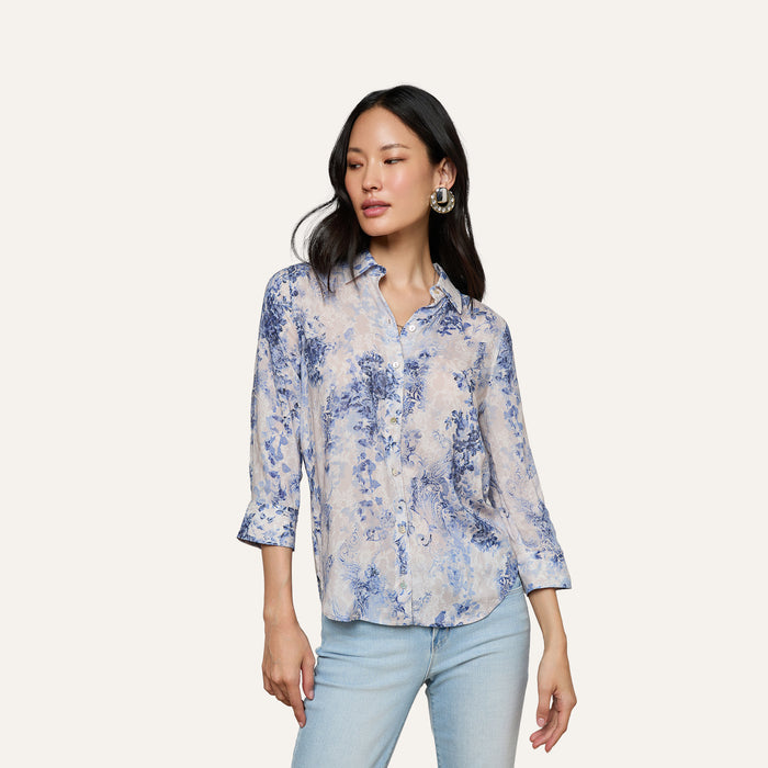 A woman stands against a plain background, one hand by her side and the other slightly raised, wearing the Camille Blouse—a light, three-quarter sleeve top crafted from viscose twill with an ivory and blue lace print—paired with light blue jeans.