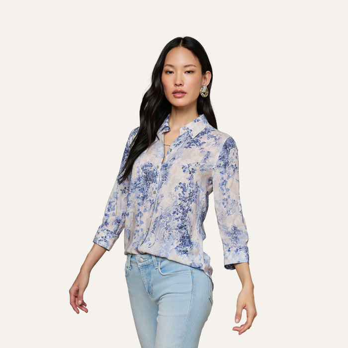 A woman wearing the Camille Blouse—a light blue and white floral button-up shirt with three-quarter sleeves, crafted from viscose twill featuring an ivory and blue lace print—paired with light blue jeans, stands against a plain background.