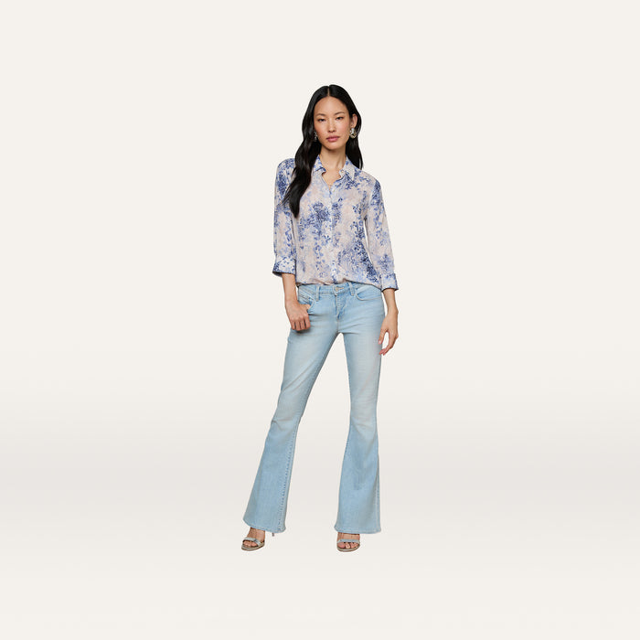 Woman standing against a plain background, wearing the Camille Blouse—an ivory and blue lace print three-quarter sleeve shirt crafted from viscose twill—paired with light-wash flared jeans and open-toe heels. She has long dark hair and a relaxed, confident pose.
