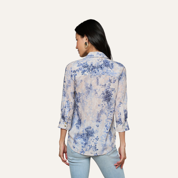 A woman with long black hair is seen from the back, wearing the Camille Blouse—an ivory and blue lace print three-quarter sleeve shirt crafted from viscose twill—paired with light blue jeans, set against a plain light background.