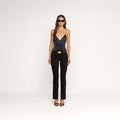 A woman wearing sunglasses, a black satin camisole, the Cara Low-Rise Bootcut Jean with a belt, and black heels stands against a plain white background.