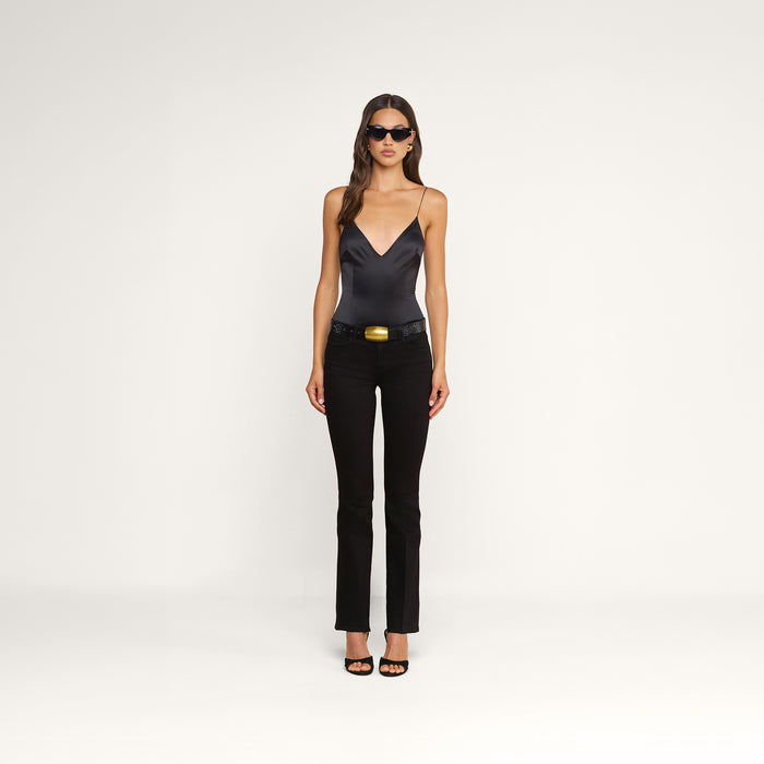 A woman wearing sunglasses, a black satin camisole, the Cara Low-Rise Bootcut Jean with a belt, and black heels stands against a plain white background.