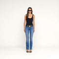 A woman stands facing forward against a plain white background, wearing sunglasses, a black tank top, the Cara Low-Rise Bootcut Jean with a belt, and black heels.