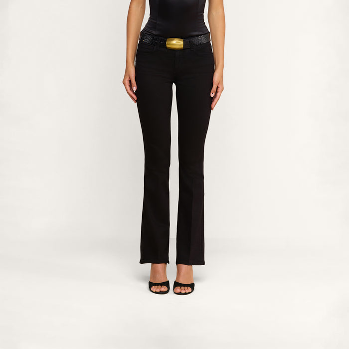 A person standing against a plain background, wearing the Cara Low-Rise Bootcut Jean in black stretch denim, paired with a black belt featuring a gold buckle and black open-toe heels. The image is cropped to show only the body from the shoulders down.