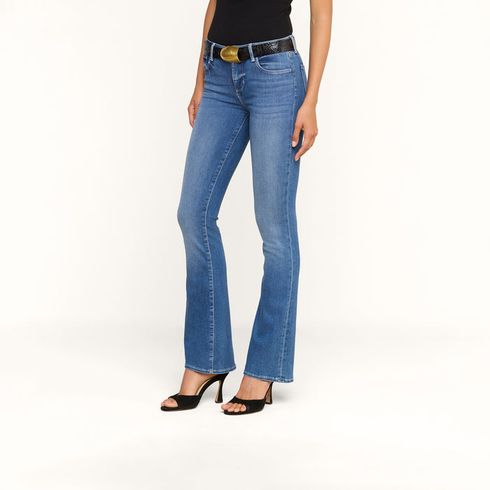 A woman stands against a plain white background wearing the Cara Low-Rise Bootcut Jean in light blue stretch denim, paired with a black belt featuring a large buckle, a black top, and black high-heeled sandals.