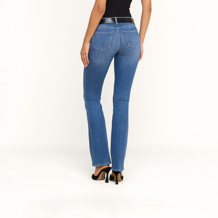 A woman wearing the Cara Low-Rise Bootcut Jean in blue stretch denim, paired with a black belt, black top, and black heeled sandals, is standing with her back to the camera against a plain white background.