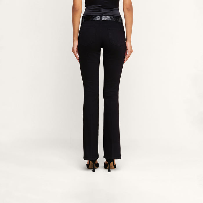 A person stands facing away, wearing the Cara Low-Rise Bootcut Jean in black stretch denim, paired with a black belt, a sleeveless black top, and black heeled shoes against a plain white background.